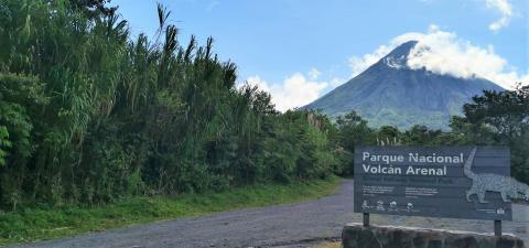 Picture of a wooden sign with the name of the park on a dirt road surrounded by leafy tropical plants and the snow capped volcano in the background 