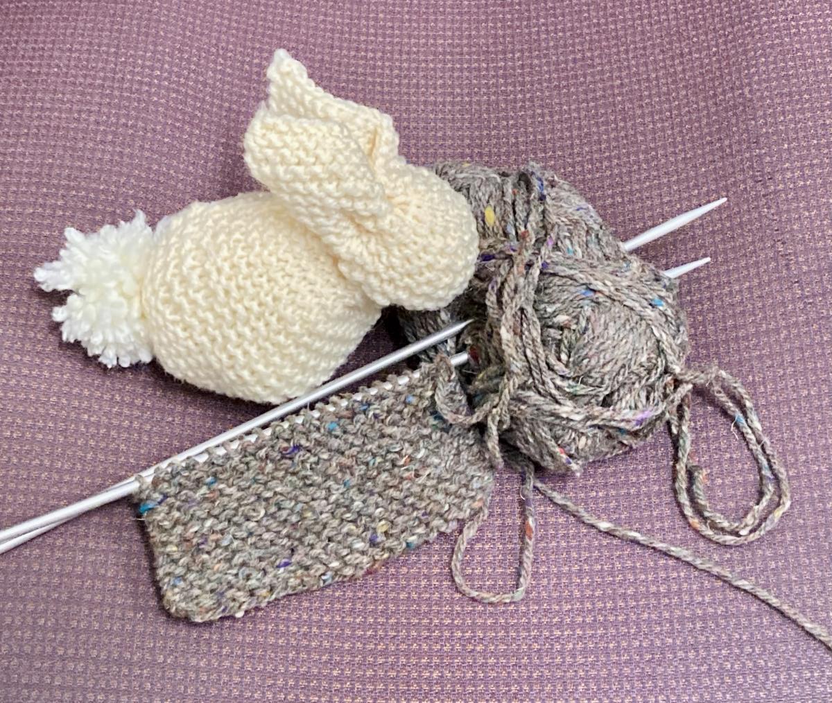 Cream colored knit bunny sits next to a project on knitting needles stabbed into gray yarn.